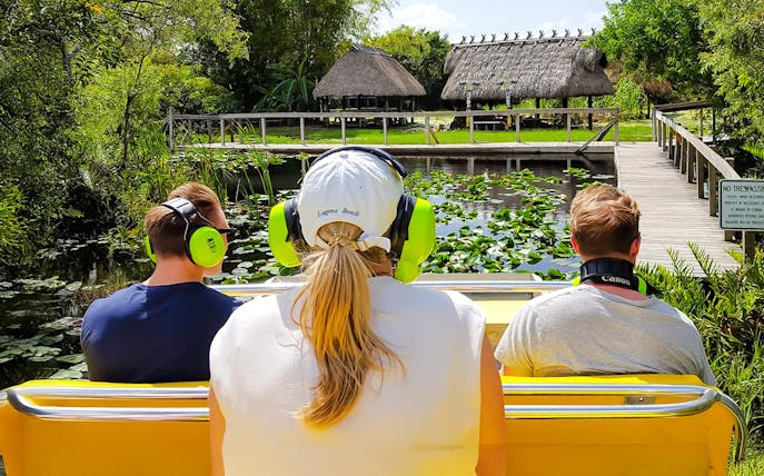 Guests on airboat ride in Everglades, viewing lush greenery and thatched huts.