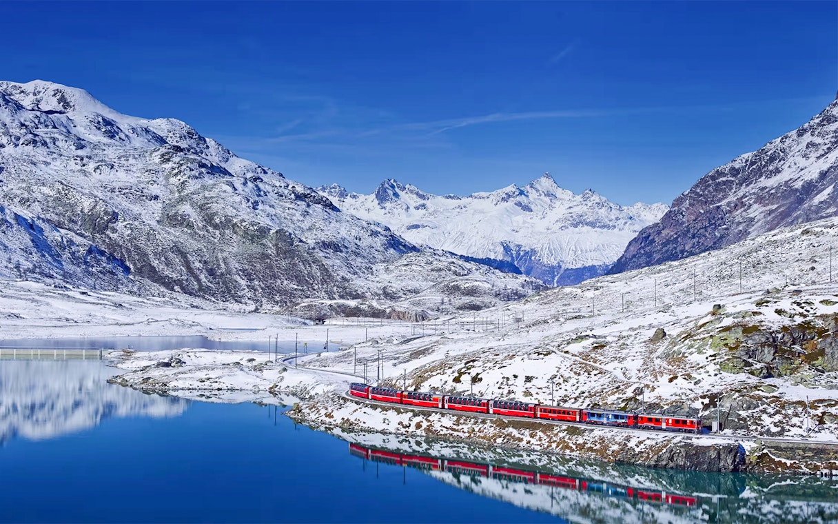Bernina Express train traveling through snowy Swiss Alps with mountain reflection in lake.