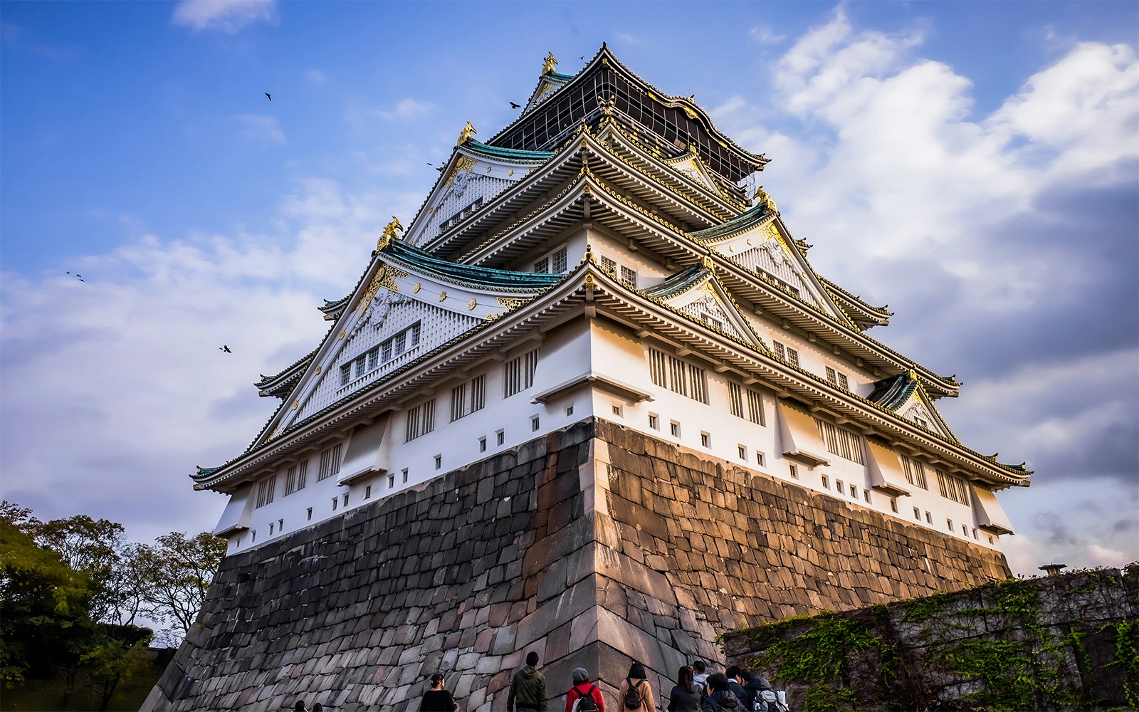 Osaka Castle with visitors approaching, set against a cloudy sky.