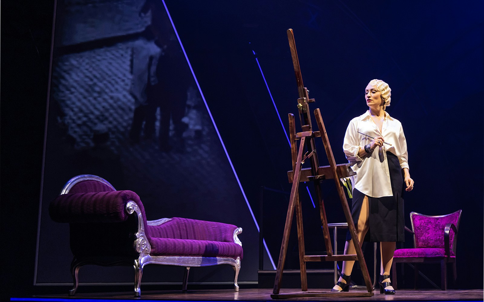 Artist painting on stage with easel and vintage furniture, Lempicka tour.