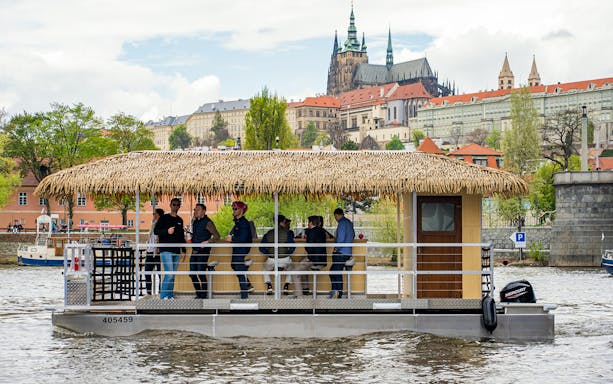 Prague Tiki Boat with people enjoying drinks, view of Prague Castle in background.
