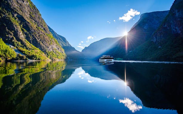 Boat cruising through Naeroyfjorden Fjord with sunlit mountains and clear reflections.