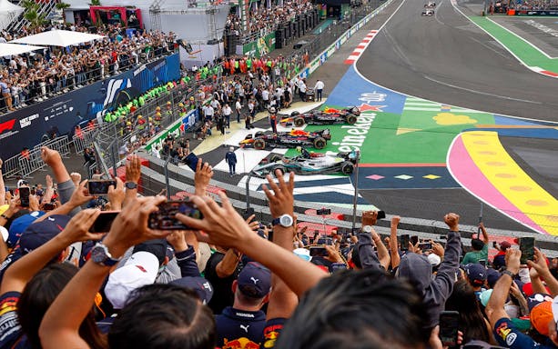 Crowd cheering at Formula 1 Mexico Grand Prix with race cars on track.