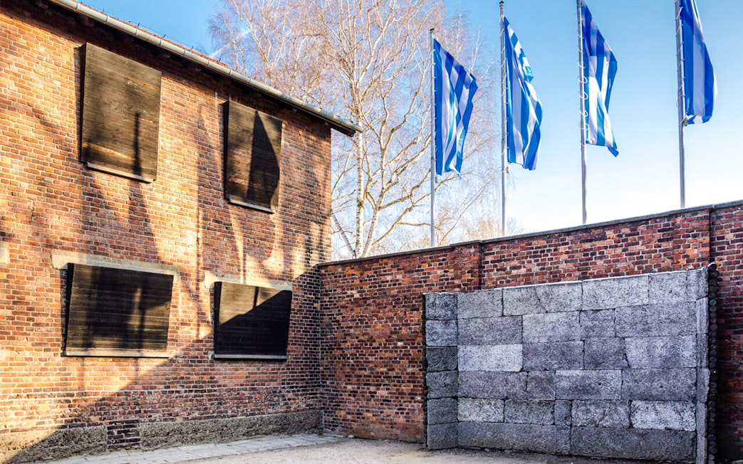 Memorial wall at Auschwitz-Birkenau Camp with brick building and flags in the background.