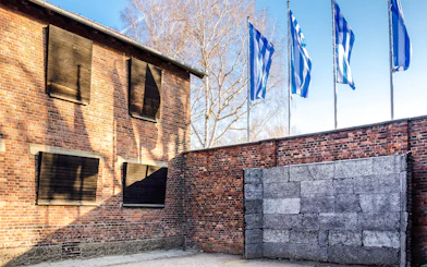 Memorial wall at Auschwitz-Birkenau Camp with brick building and flags in the background.