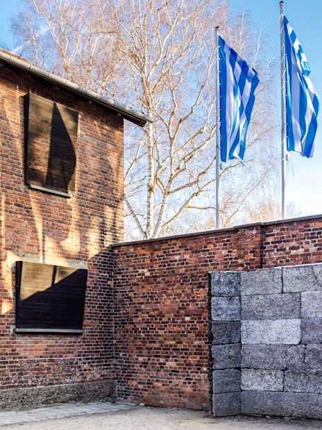 Memorial wall at Auschwitz-Birkenau Camp with brick building and flags in the background.