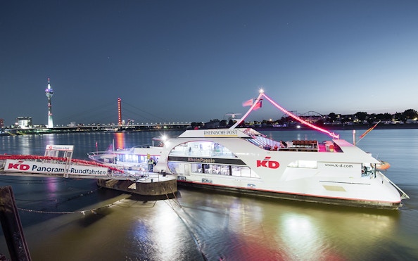 Evening cruise ship on the Rhine River with Düsseldorf skyline and Rheinturm in the background.