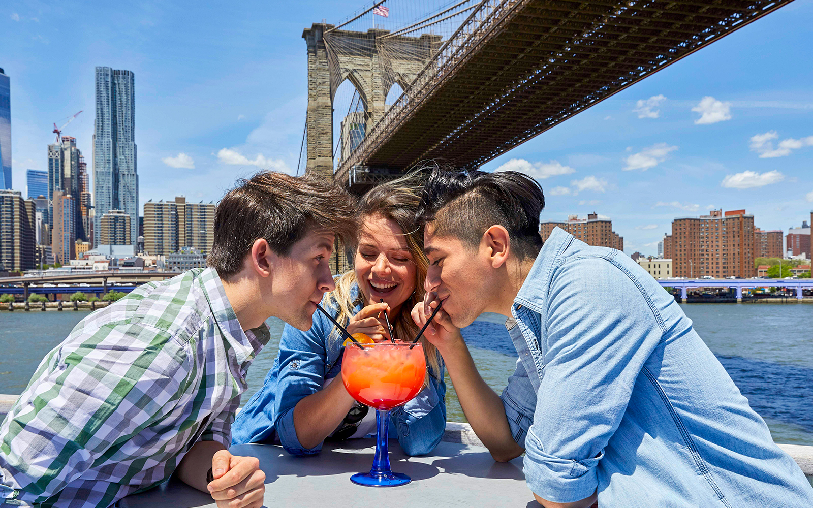 Friends sharing a drink on a New York dinner cruise with Brooklyn Bridge in the background.