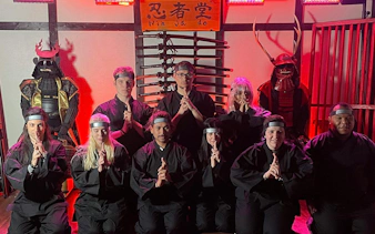 Participants in ninja attire at Osaka Ninja Workshop with samurai armor display.