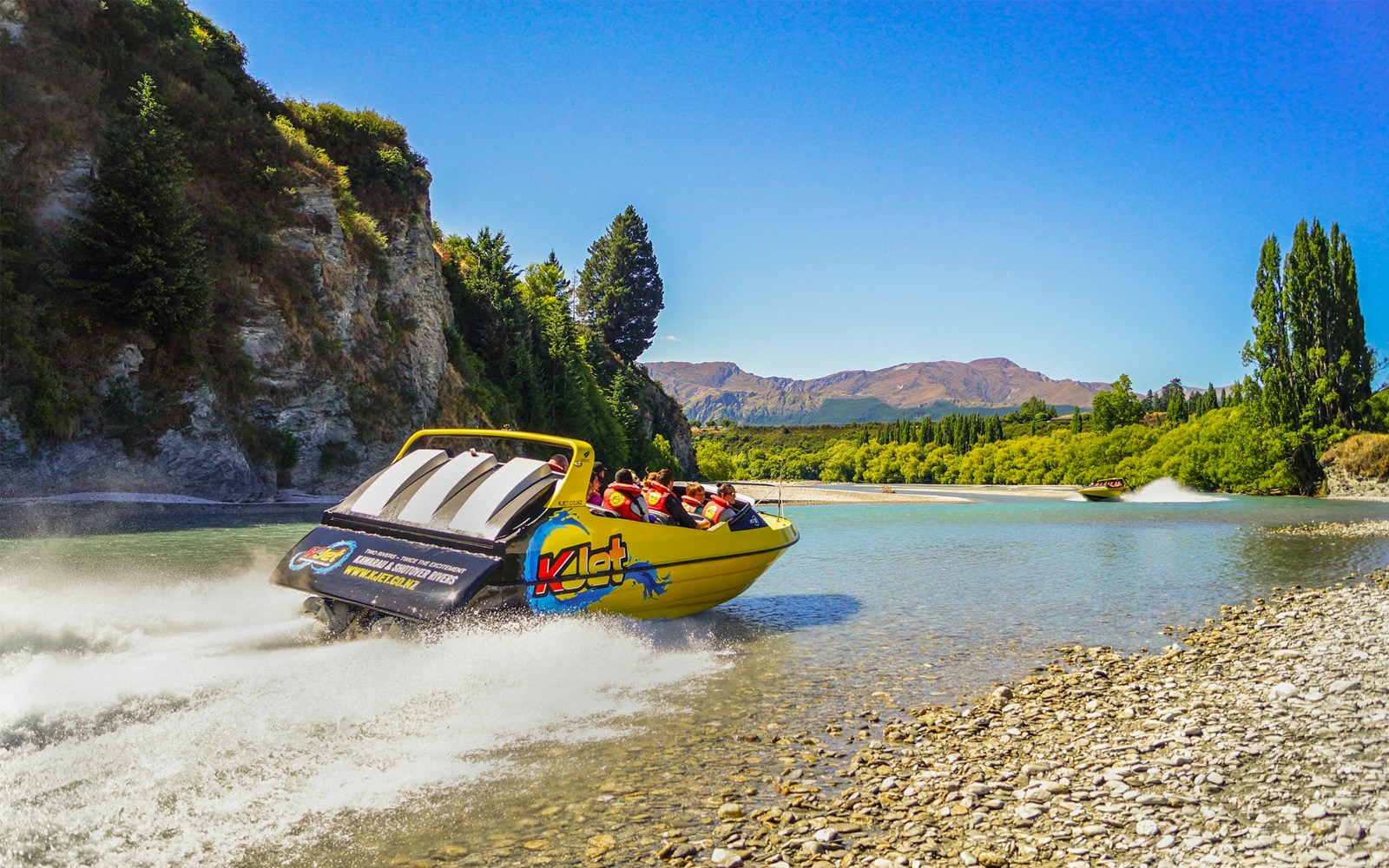 Jet boat speeding on Kawarau River with passengers, surrounded by scenic cliffs and trees.