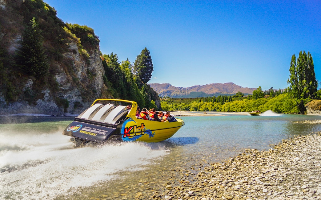 Jet boat speeding on Kawarau River with passengers, surrounded by scenic cliffs and trees.