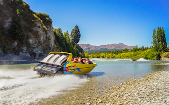 Jet boat speeding on Kawarau River with passengers, surrounded by scenic cliffs and trees.