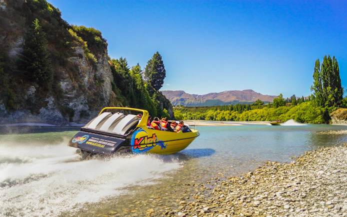 Jet boat speeding on Kawarau River with passengers, surrounded by scenic cliffs and trees.