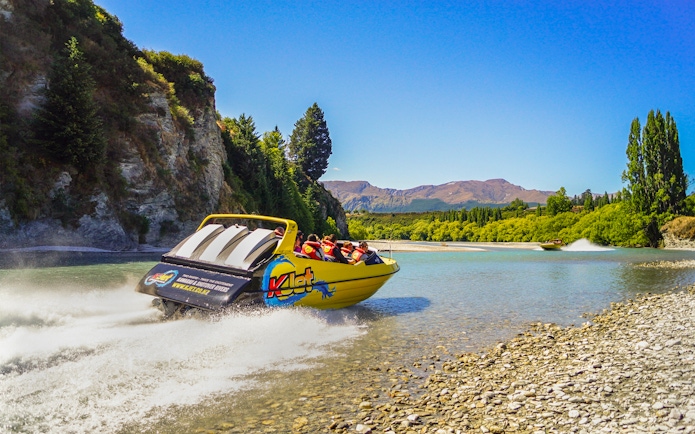 Jet boat speeding on Kawarau River with passengers, surrounded by scenic cliffs and trees.