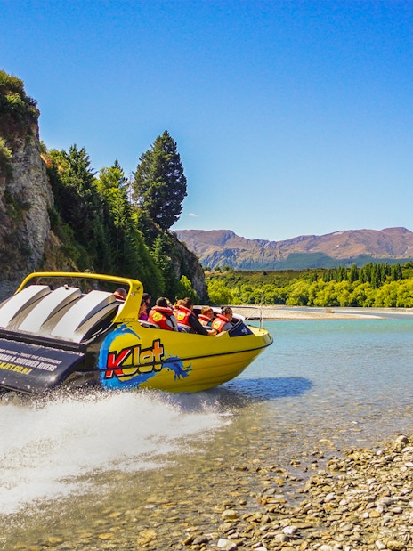 Jet boat speeding on Kawarau River with passengers, surrounded by scenic cliffs and trees.
