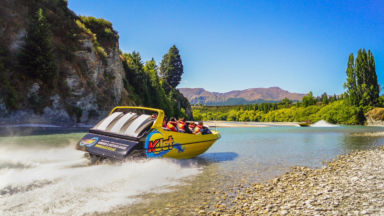 Jet boat speeding on Kawarau River with passengers, surrounded by scenic cliffs and trees.