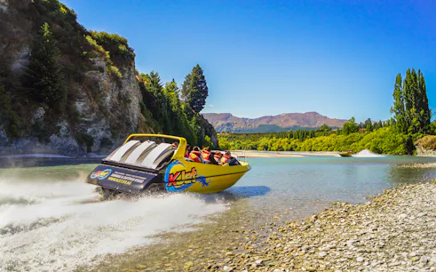 Jet boat speeding on Kawarau River with passengers, surrounded by scenic cliffs and trees.