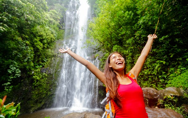 Woman smiling with arms raised in front of waterfall, Road to Hana, Hawaii.