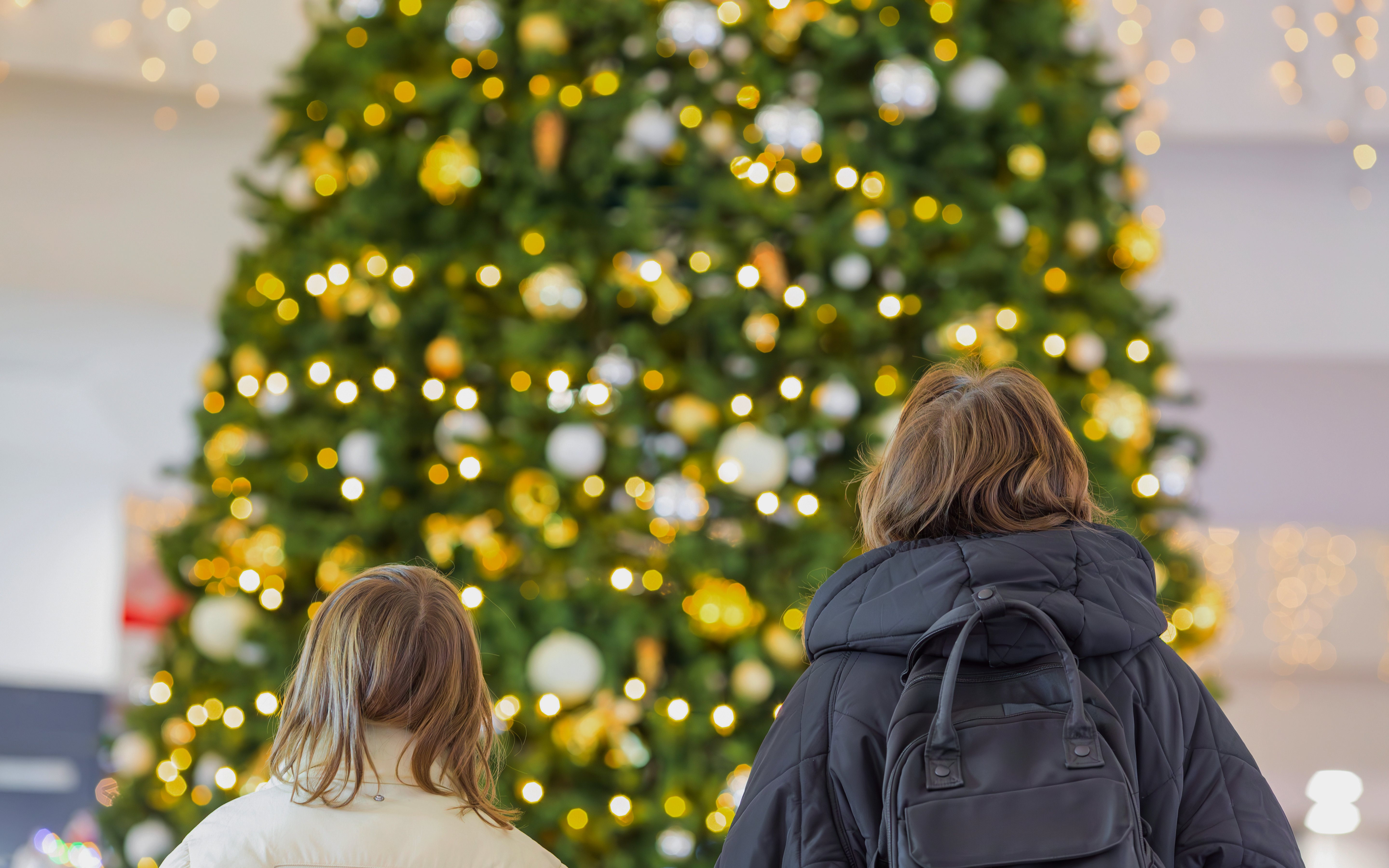 Shoppers admiring a large decorated Christmas tree with lights in a mall.