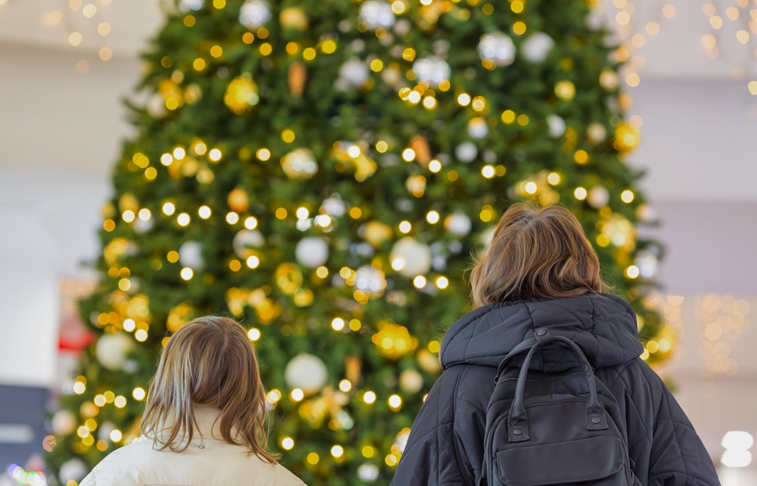 Shoppers admiring a large decorated Christmas tree with lights in a mall.