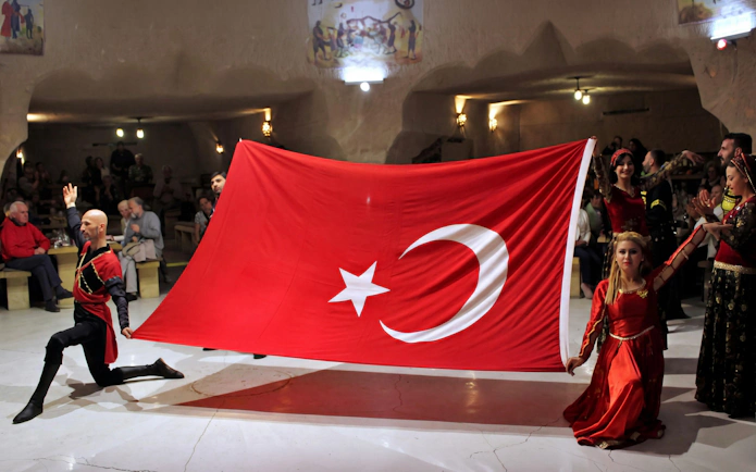 Traditional Turkish folk dancers performing with a large Turkish flag indoors.