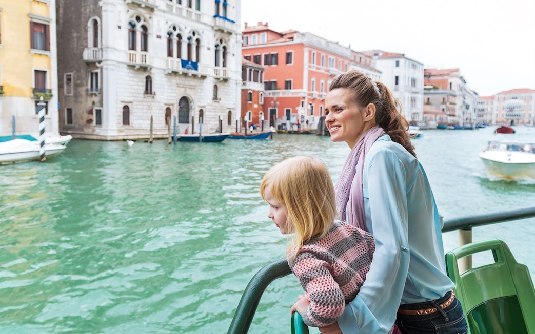 Mother and daughter on a water bus in Venice, with historic buildings in the background.