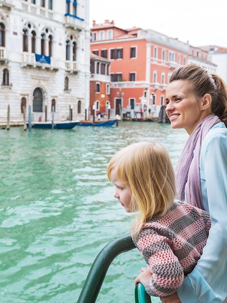 Mother and daughter on a water bus in Venice, with historic buildings in the background.