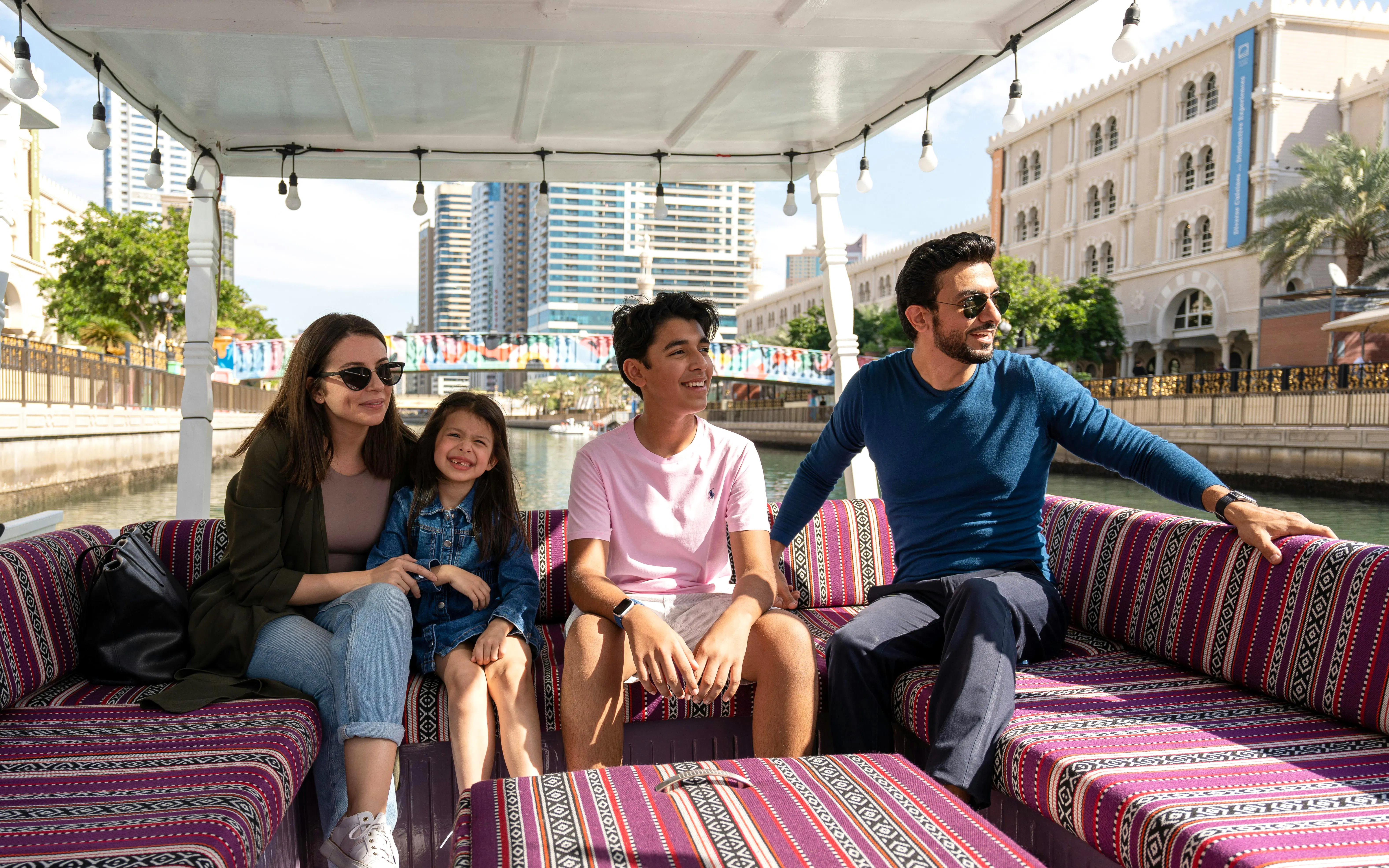Family enjoying a boat tour on a canal in Sharjah with cityscape views.