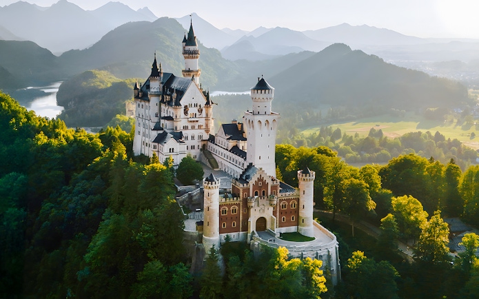 Aerial view of Neuschwanstein Castle surrounded by lush green forests and mountains.