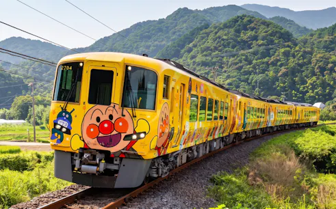 Colorful train with cartoon characters on JR PASS Kagawa Mini Rail, surrounded by lush green hills.