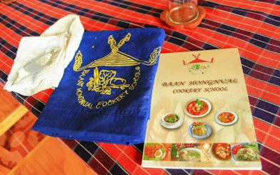 Baan Hongnual Cookery School brochure and apron on a checkered tablecloth in Chiang Mai.
