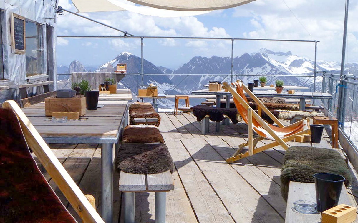 Outdoor seating area with wooden tables and chairs overlooking snowy mountains at Glacier 3000.