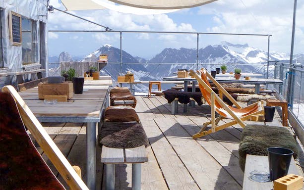 Outdoor seating area with wooden tables and chairs overlooking snowy mountains at Glacier 3000.