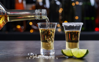 Tequila being poured into a shot glass on a bar counter with lime wedges.