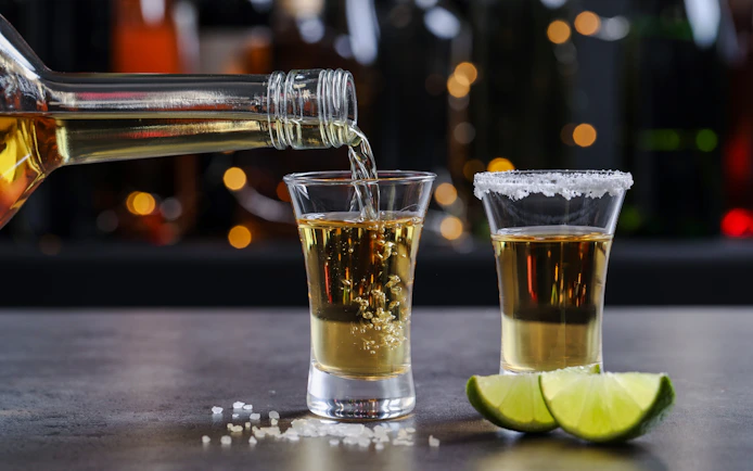 Tequila being poured into a shot glass on a bar counter with lime wedges.