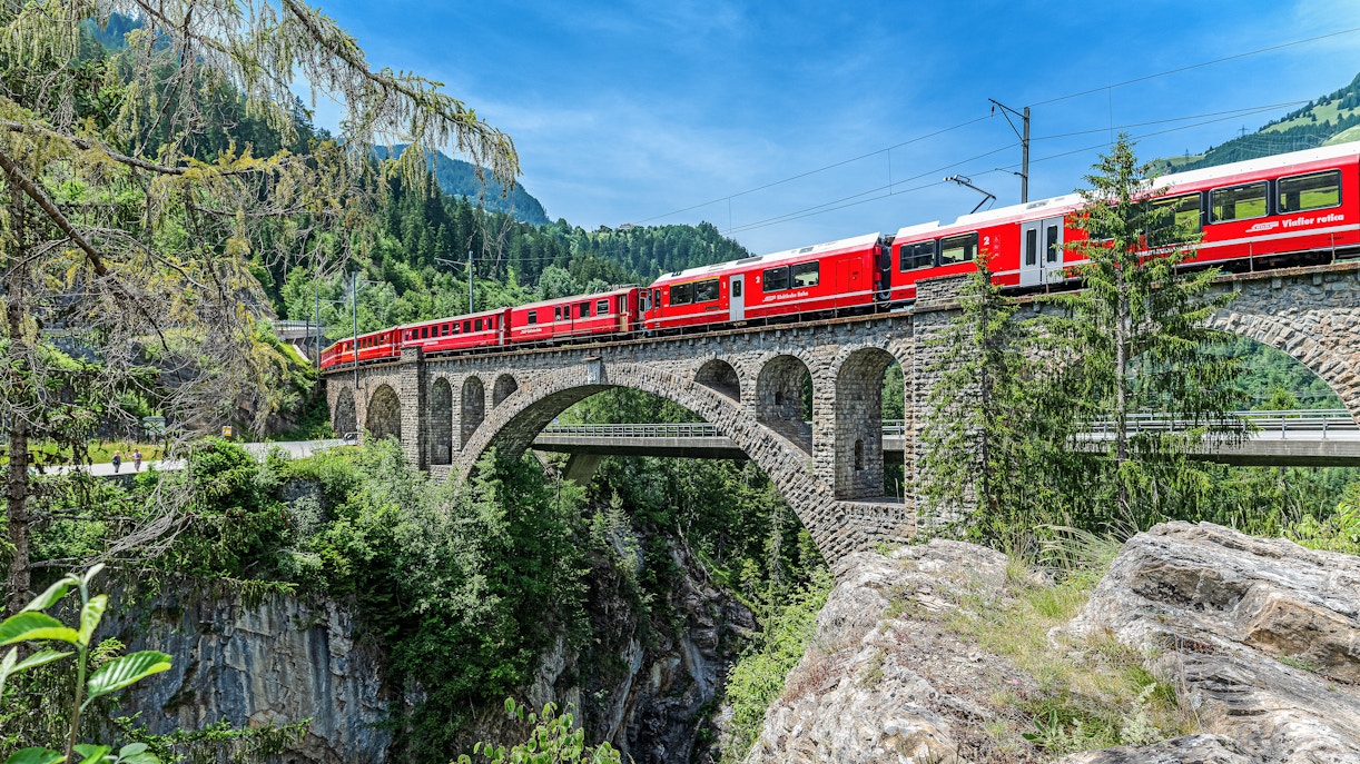 Bernina train crossing Solis Viaduct in Swiss Alps with lush greenery and rocky landscape.