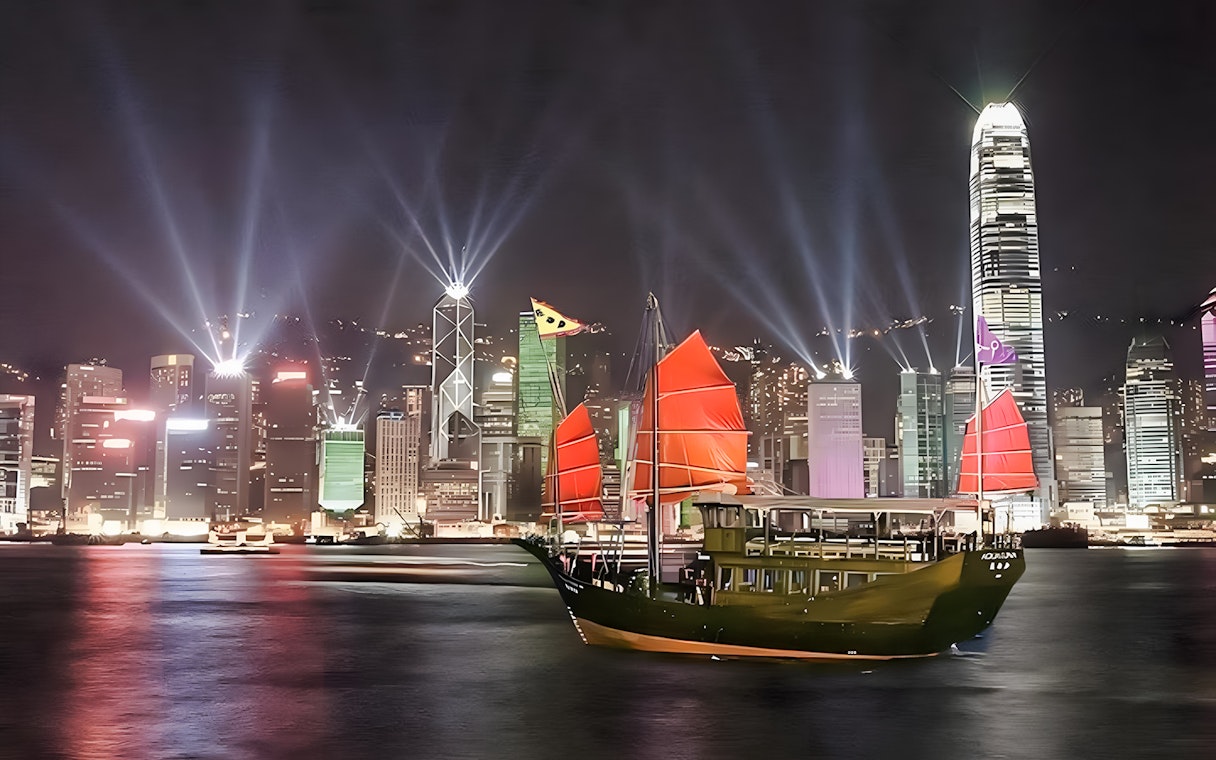 Aqua Luna cruise with red sails in Hong Kong harbor at night, city skyline illuminated.