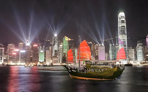 Aqua Luna cruise with red sails in Hong Kong harbor at night, city skyline illuminated.