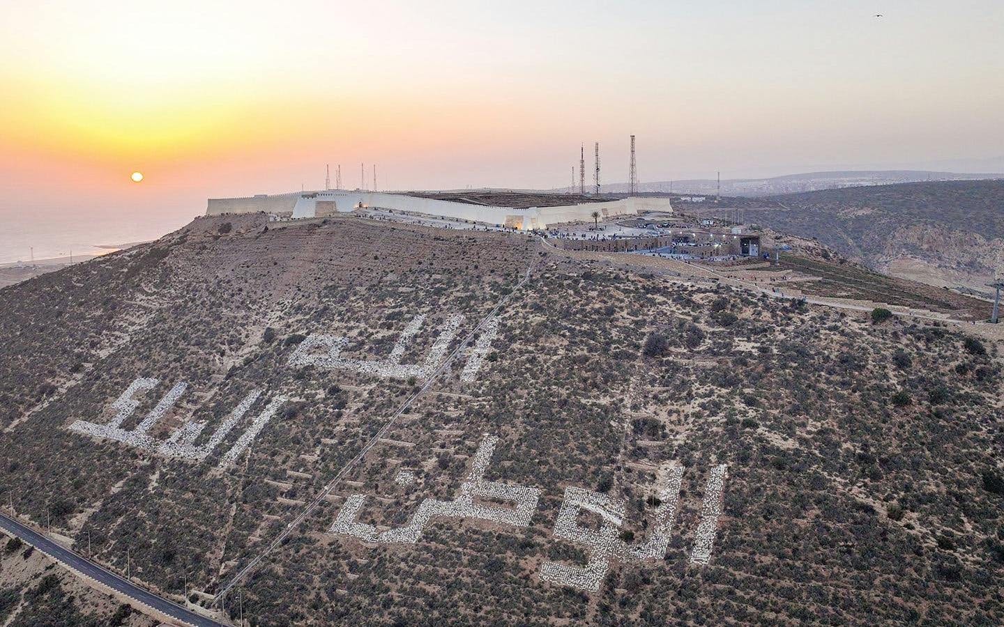 Hilltop view of Agadir with Arabic inscription at sunset, featuring cityscape and coastline.