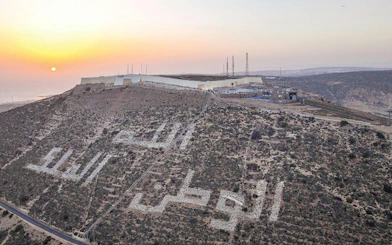 Hilltop view of Agadir with Arabic inscription at sunset, featuring cityscape and coastline.