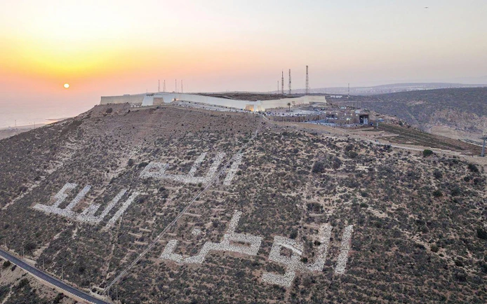 Hilltop view of Agadir with Arabic inscription at sunset, featuring cityscape and coastline.