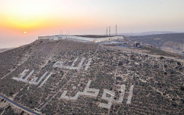 Hilltop view of Agadir with Arabic inscription at sunset, featuring cityscape and coastline.