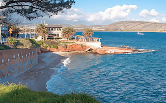 Coastal view of Sounion with a sandy beach and a building surrounded by palm trees.
