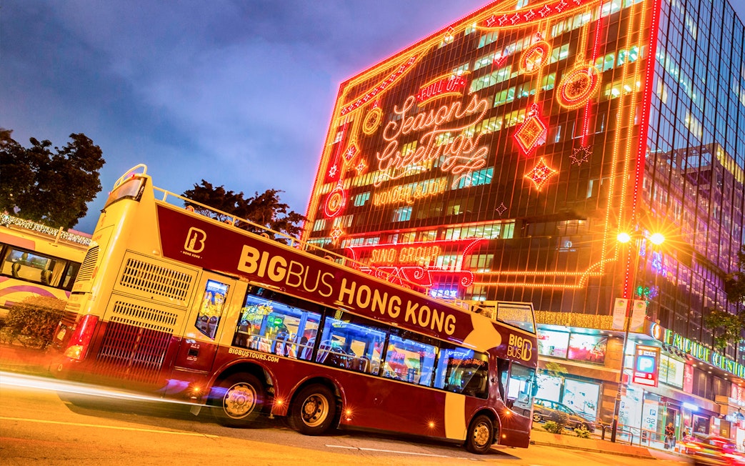 Hop-on-hop-off bus in Hong Kong with festive lights on a building.