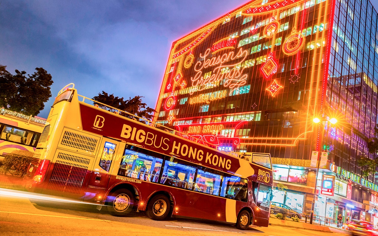 Hop-on-hop-off bus in Hong Kong with festive lights on a building.