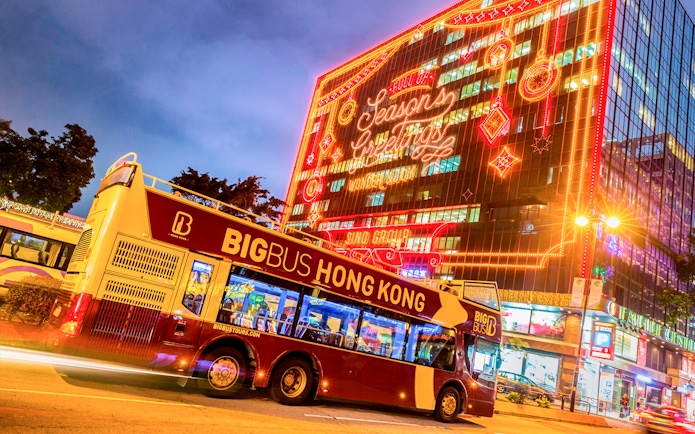 Hop-on-hop-off bus in Hong Kong with festive lights on a building.