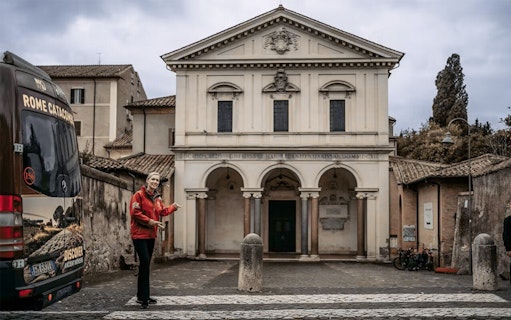 Tour guide near entrance of Rome catacombs with tour bus in foreground.