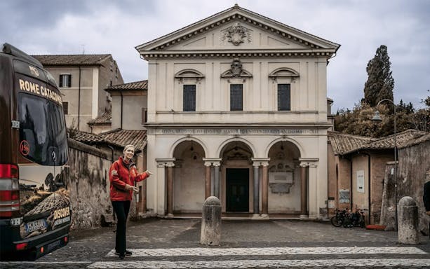 Tour guide near entrance of Rome catacombs with tour bus in foreground.