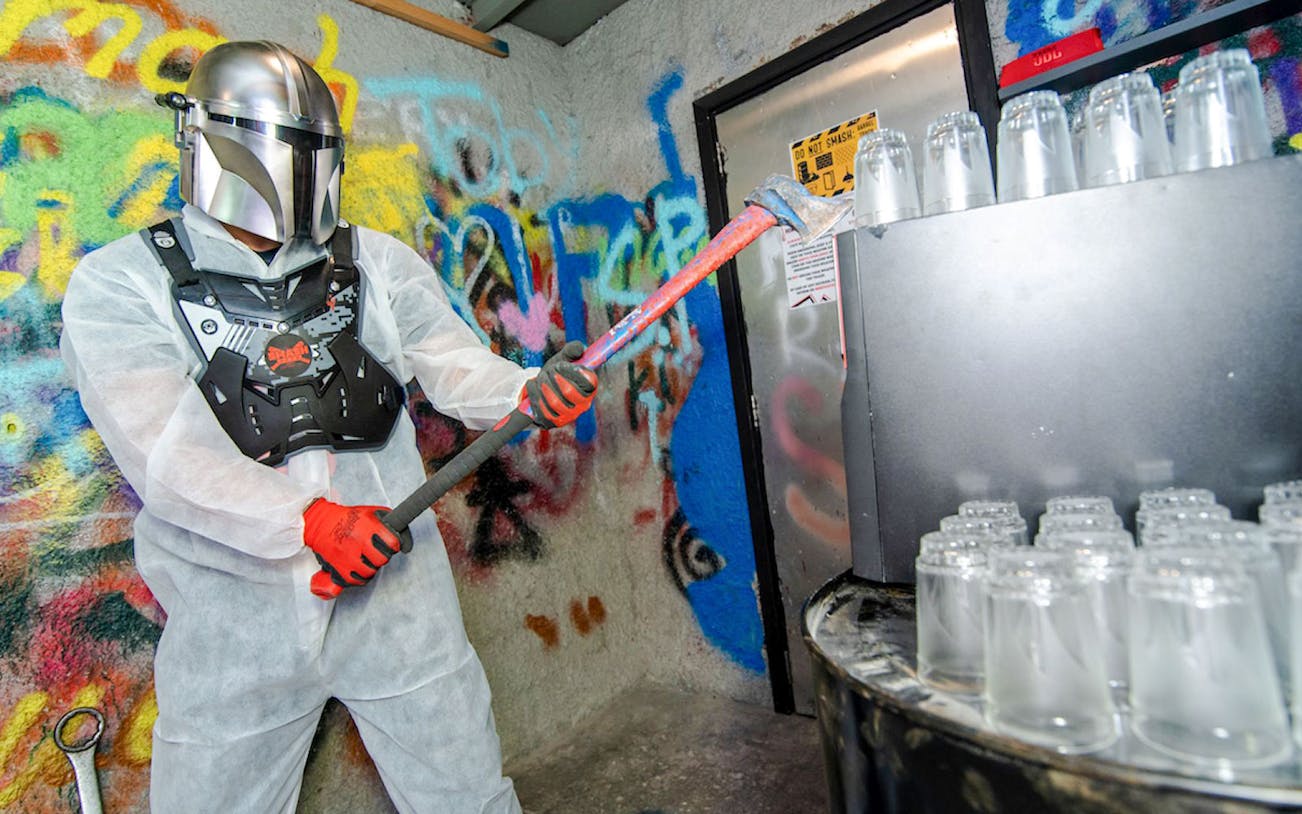 Visitor in protective gear smashing bottles in a graffiti-covered smash room.