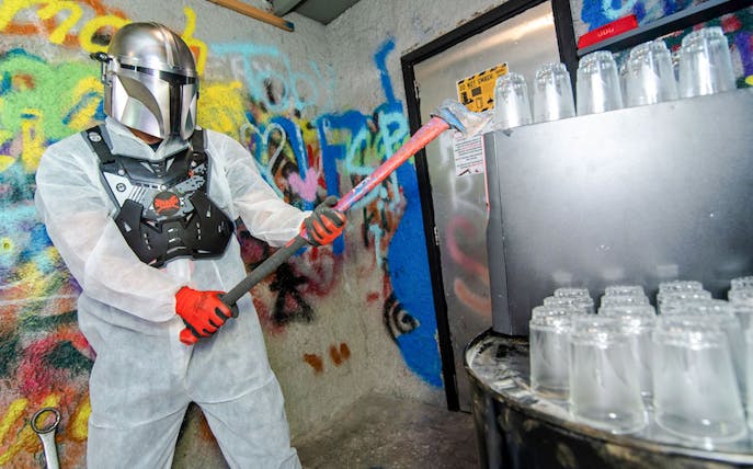Visitor in protective gear smashing bottles in a graffiti-covered smash room.